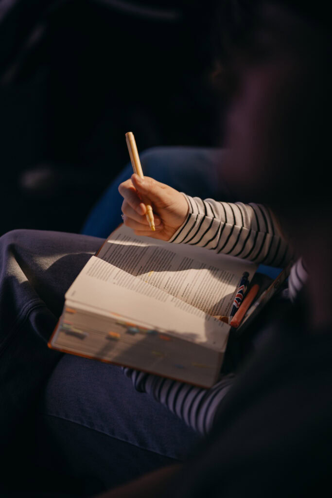 Ruka drží žlutou propisku nad otevřenou knihou na klíně osoby v pruhovaném tričku.