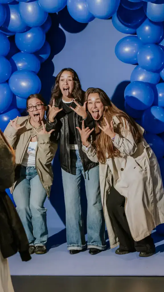 Drei Frauen posieren lachend vor einem blauen Ballonbogen.