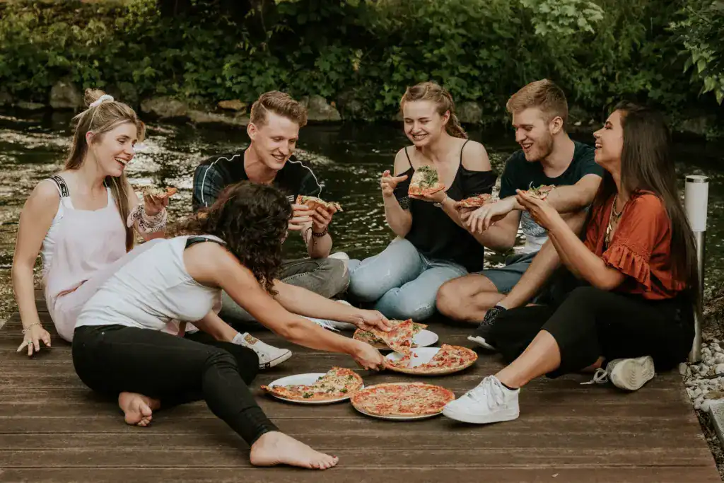Sechs junge Menschen sitzen auf einem Holzsteg am Wasser und essen Pizza.