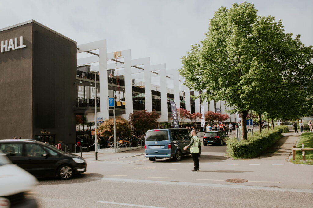Straßenansicht mit Parkhaus, Bäumen und Autos, Fußgänger auf der Straße, bei bewölktem Himmel.