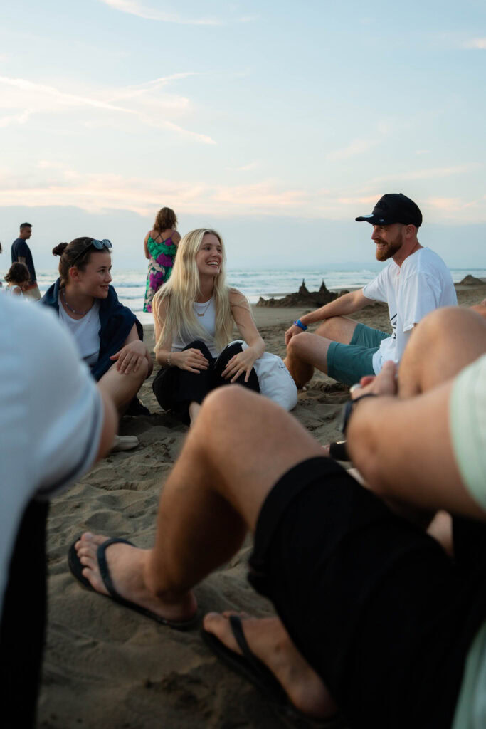 Gruppe von Menschen sitzt am Strand, unterhalten sich und lachen.