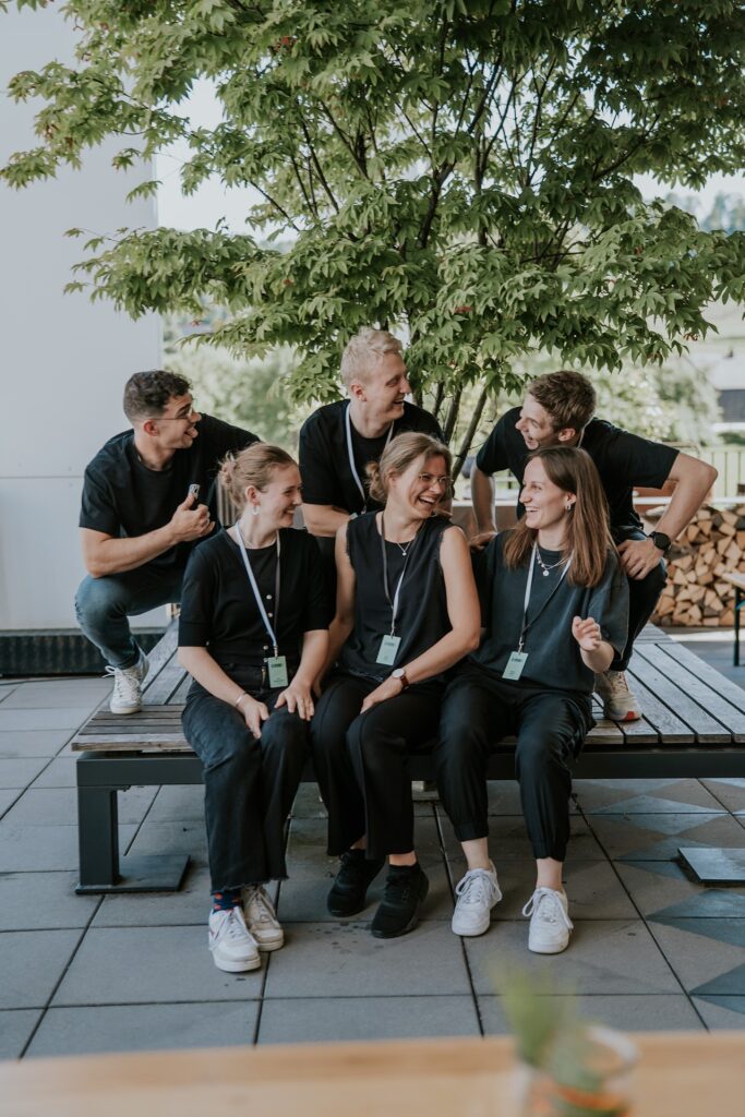 Sechs junge Menschen sitzen und stehen auf einer Terrasse unter einem Baum, lachen und sprechen.