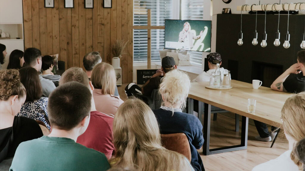 People are sitting in the room watching a TV with a person on it.