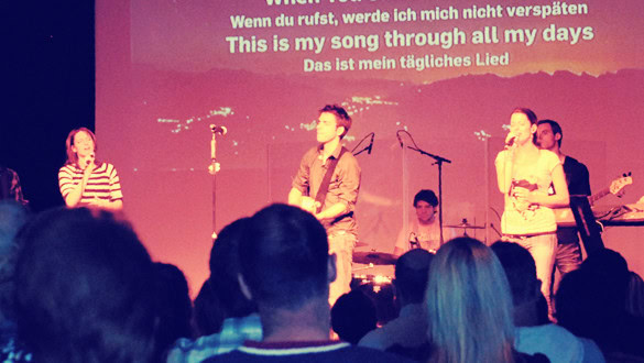 Vier Musiker auf einer Bühne mit Mikrofonen und Instrumenten, im Hintergrund Text.