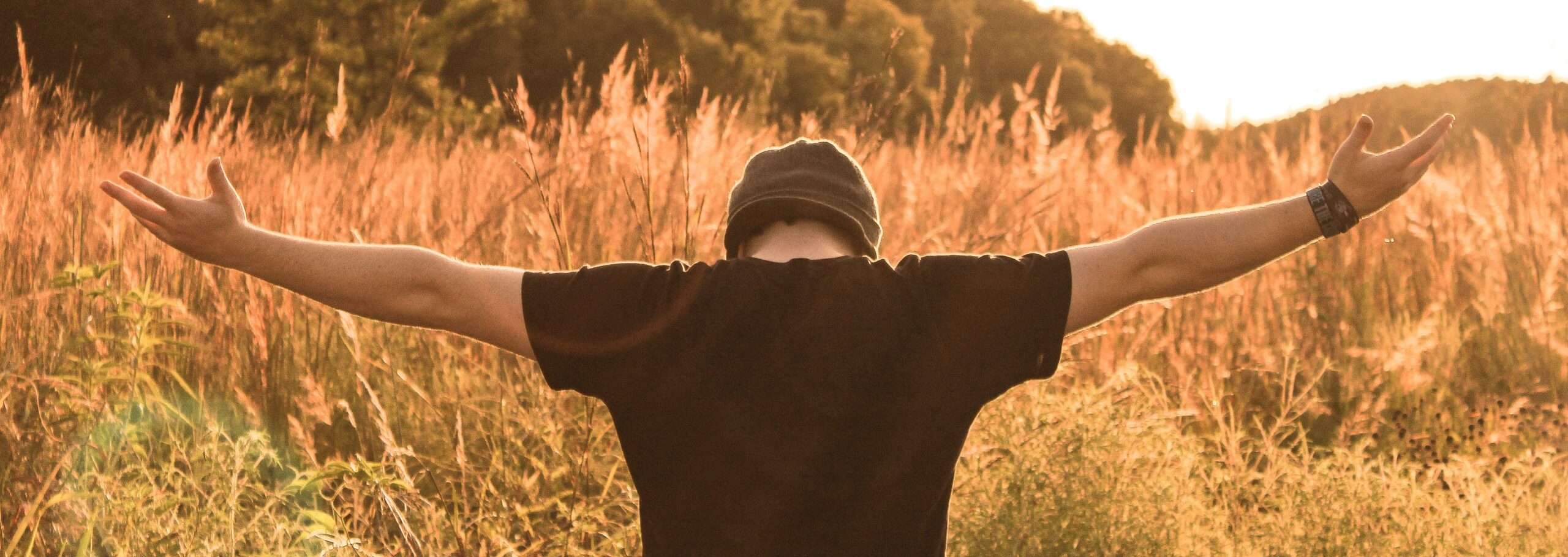 Person mit schwarzen T-Shirt und Mütze mit ausgebreiteten Armen in einem Feld bei Sonnenuntergang.