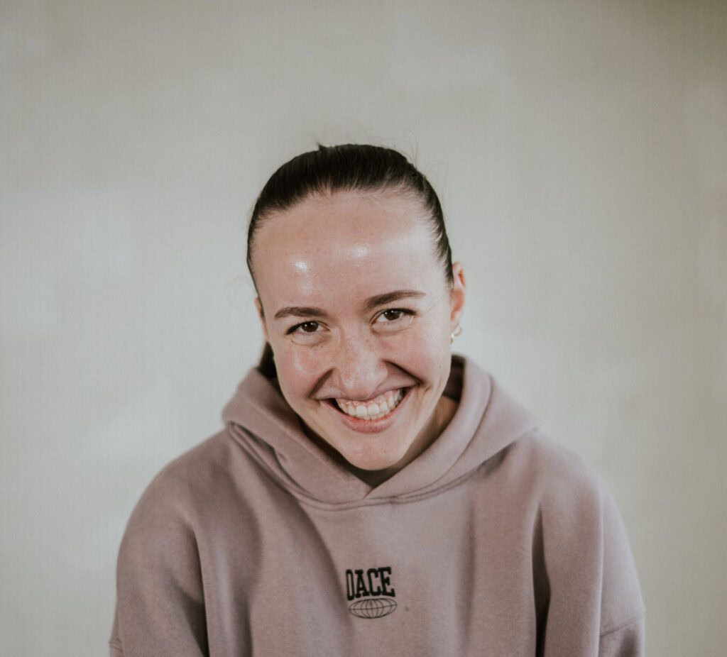 Junge Frau mit braunem Haar in einem hellbraunen Hoodie, lächelnd, vor neutralem Hintergrund.