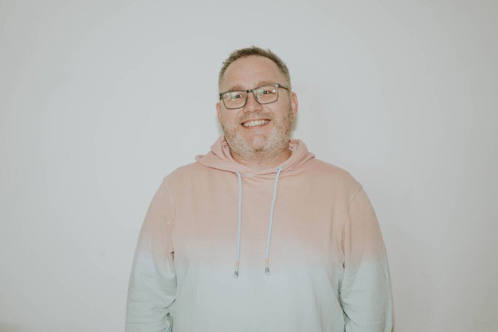 Mann mit Brille und Hoodie lächelt vor weißem Hintergrund.