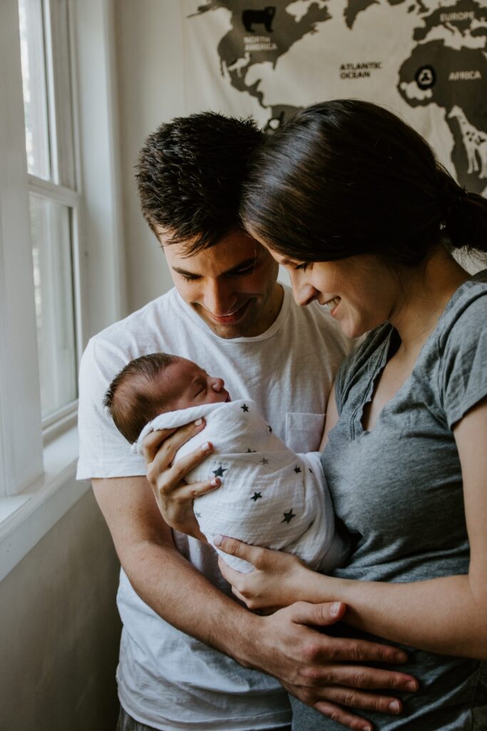 A happy couple holds their newborn baby in their arms, with a world map on the wall in the background, symbolizing global community and family.
