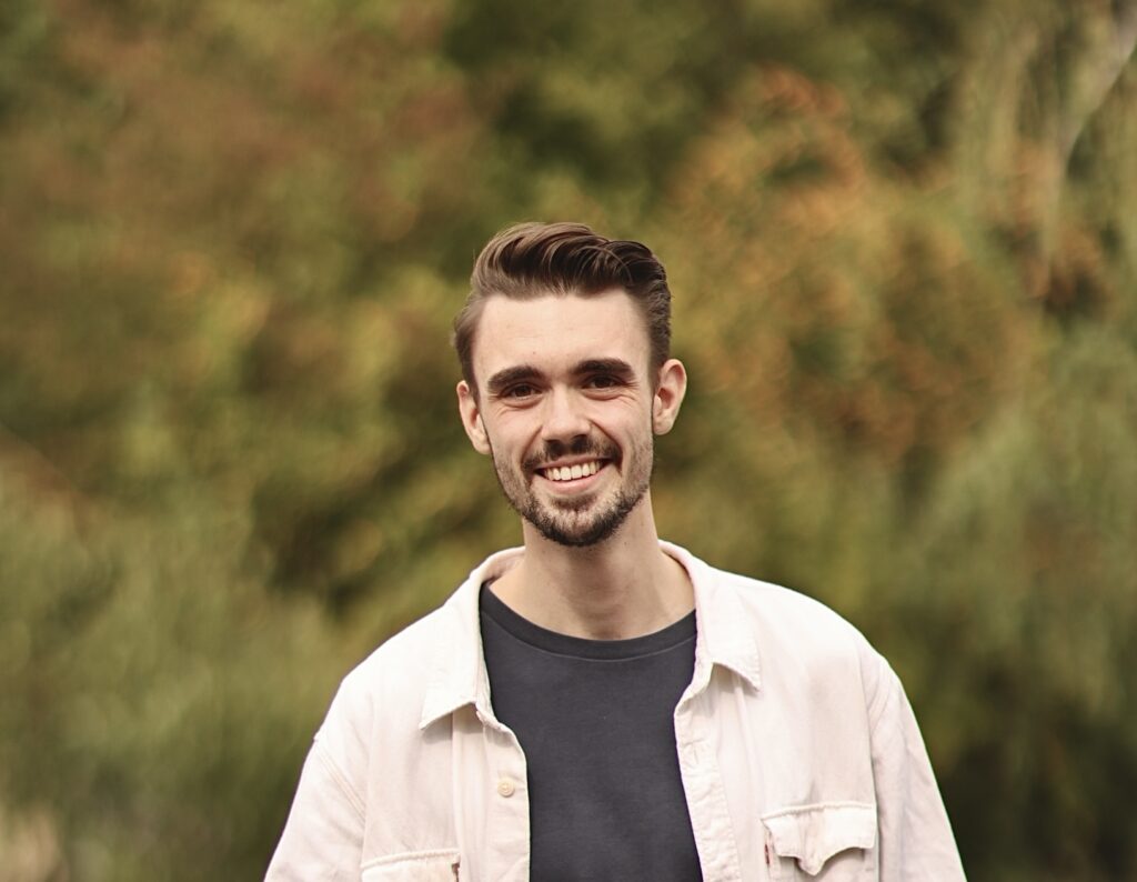 Junger Mann mit dunklem T-Shirt und heller Jacke, lächelnd im Park.