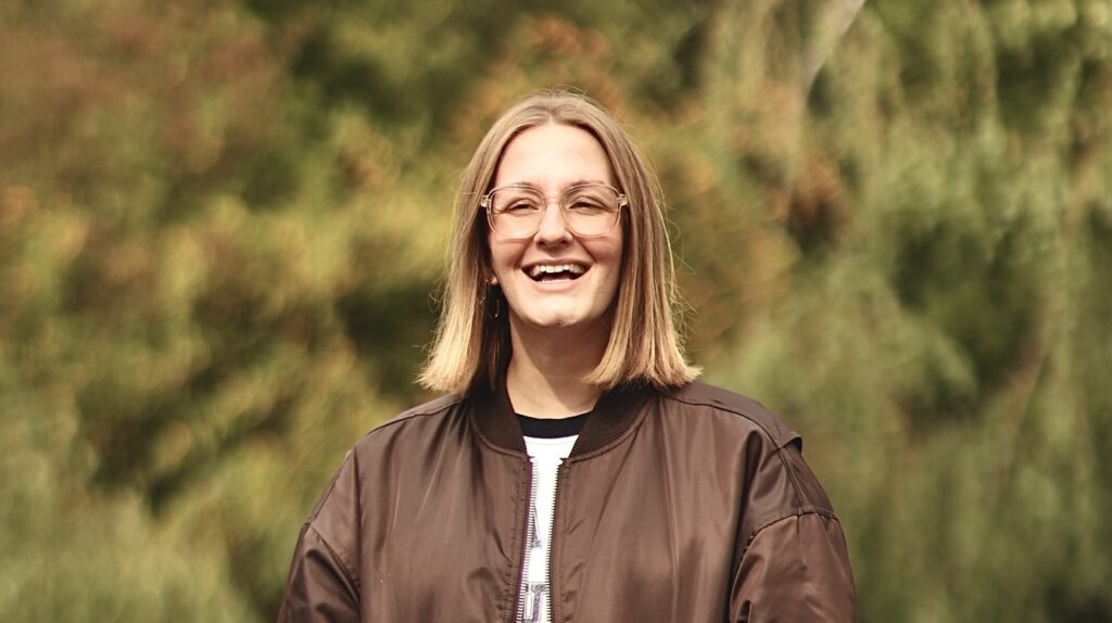 Frau mit Brille und braunem Jacke lächelt im Freien.