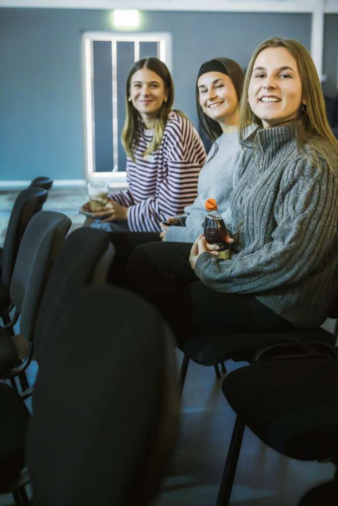 Drei junge Frauen sitzen in einem Wartezimmer, eine hält ein Getränk in der Hand.