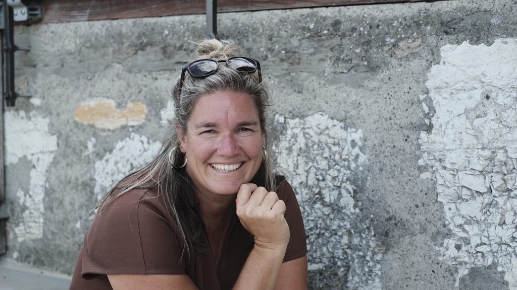 Frau mit Brille, die lächelnd vor einer alten Wand sitzt.