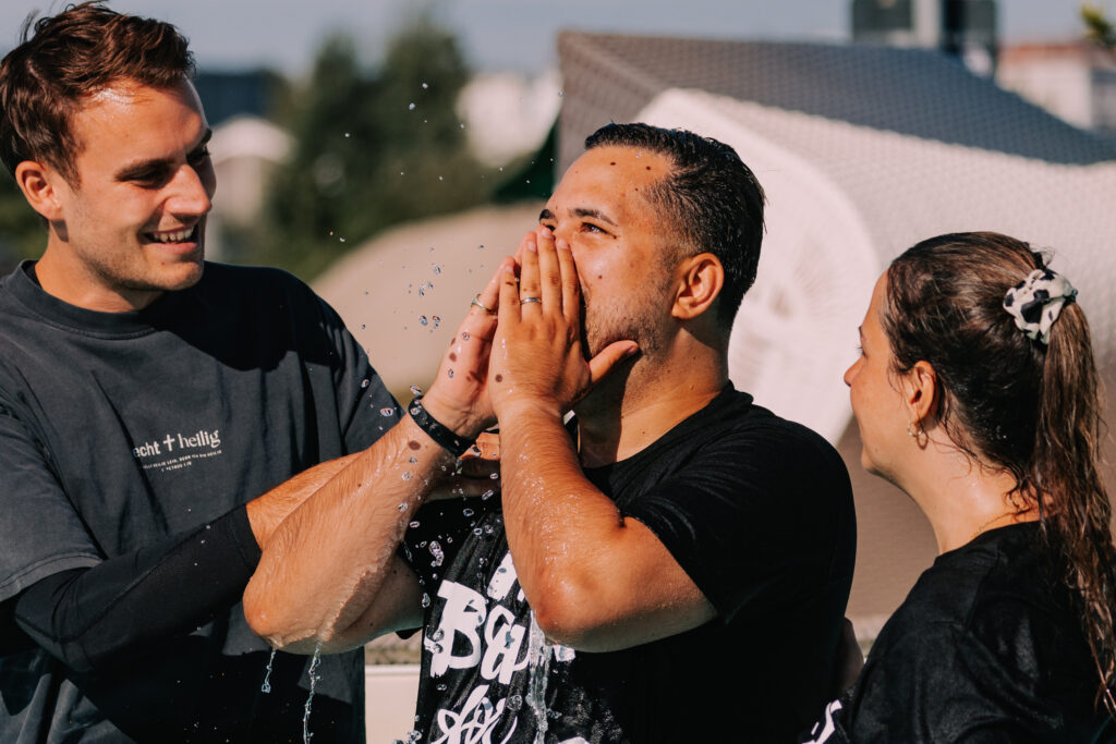 Zwei Männer und eine Frau feiern gemeinsam im Freien, Wasser wird gespritzt, Freude und Gemeinschaft im christlichen Kontext.
