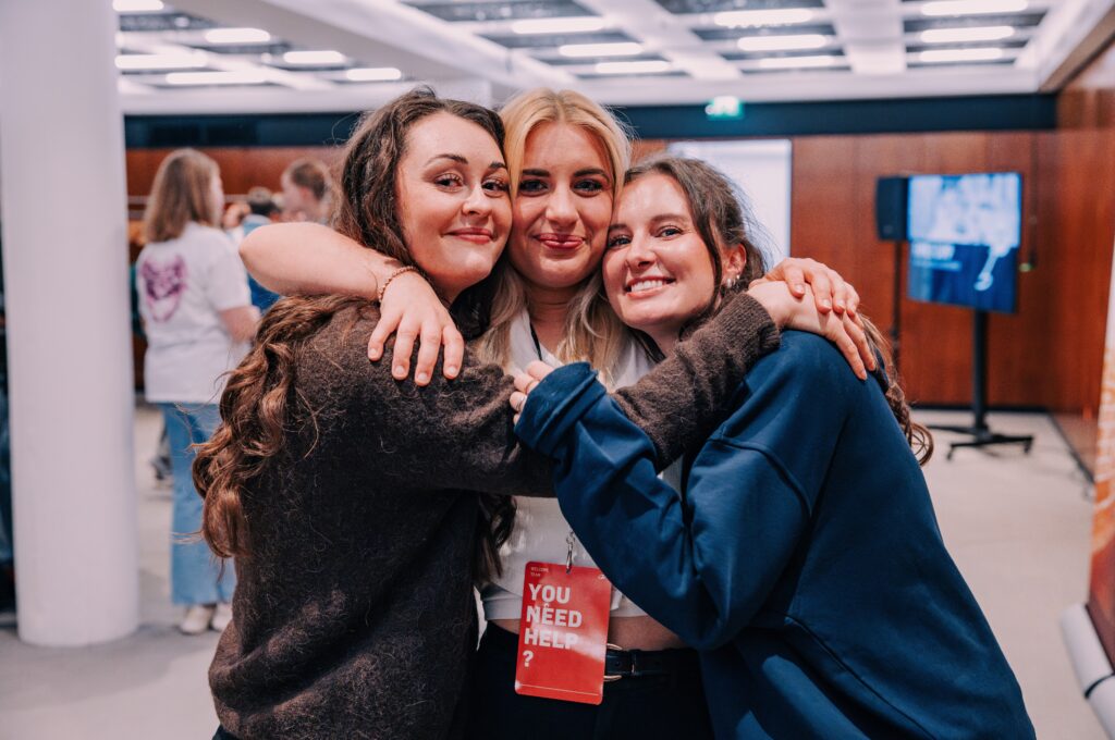 Drei lachende Frauen umarmen sich bei einem Gemeinschaftsevent im ICF Hamburg, die Atmosphäre ist herzlich und freundlich, mit weiteren Teilnehmern im Hintergrund.