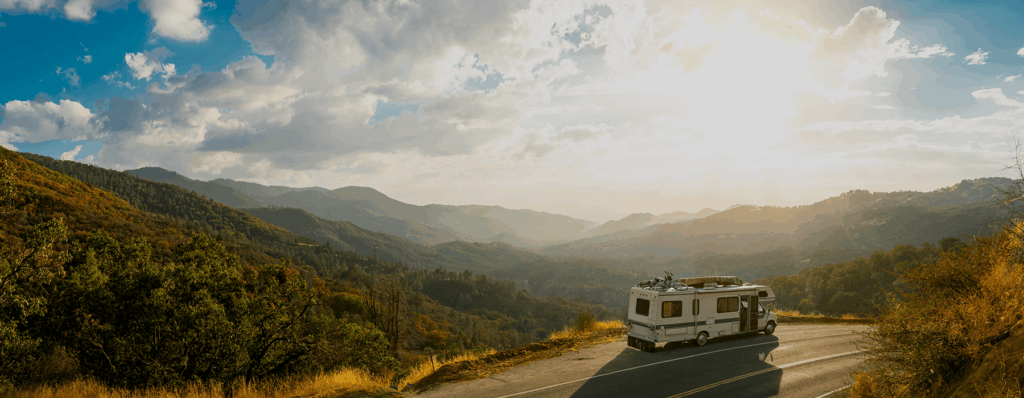 Ein Wohnmobil fährt auf einer kurvigen Straße durch eine malerische Berglandschaft bei Sonnenuntergang, umgeben von grünen Hügeln und bewölktem Himmel.