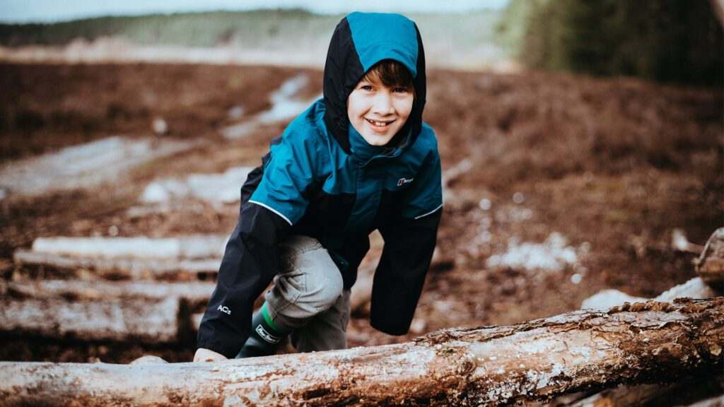 Junge Person in blauer Jacke klettert auf Baumstamm im Freien.