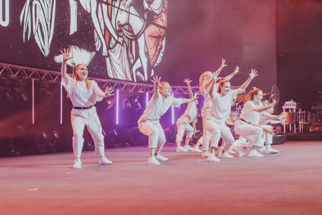 Sechs Tänzerinnen in weißen Outfits tanzen auf Bühne mit Licht und Hintergrundbild.