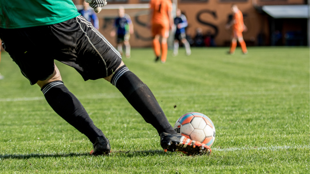 Fuß eines Fußballspielers beim Schuss auf den Ball, andere Spieler im Hintergrund.