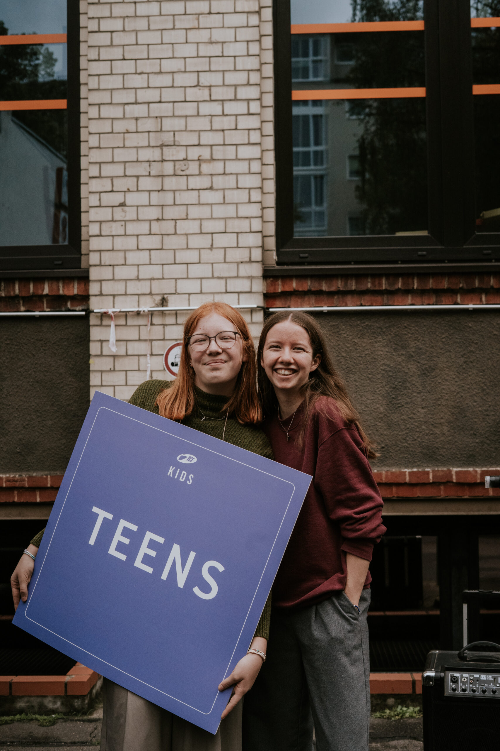 Zwei junge Frauen halten ein großes Schild mit 'TEENS' vor einem Gebäude.