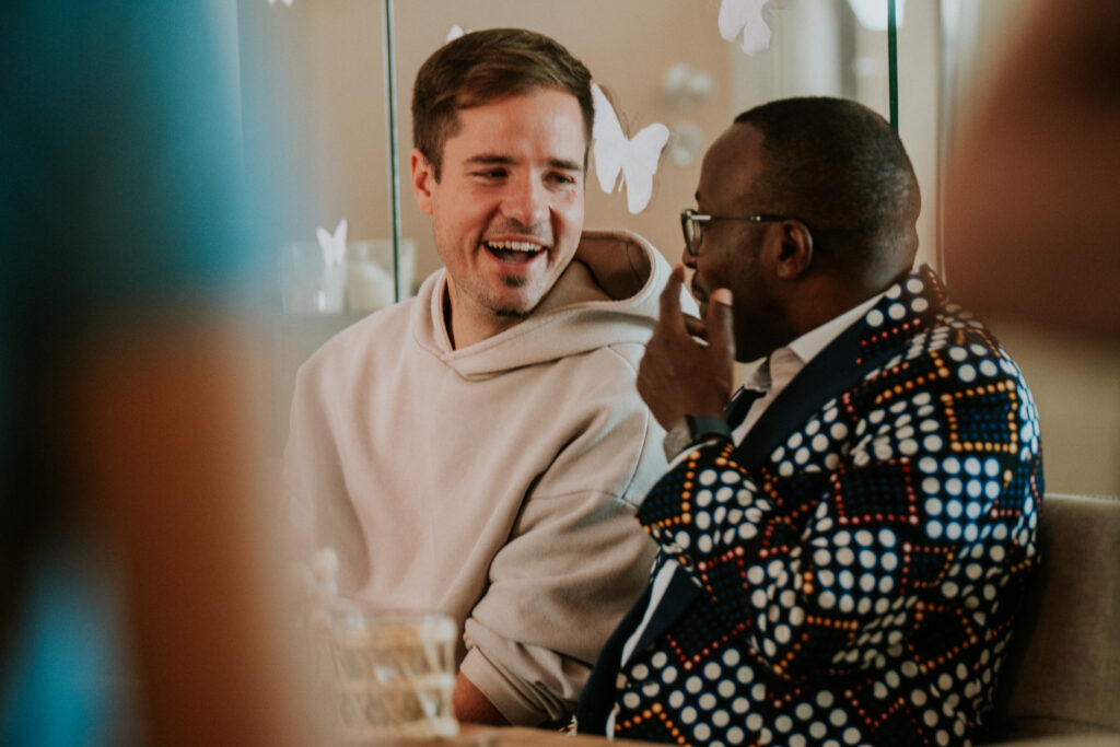 Zwei Männer sprechen lachend in einem Café, einer trägt ein helles Hoodie, der andere ein bunt gemal.