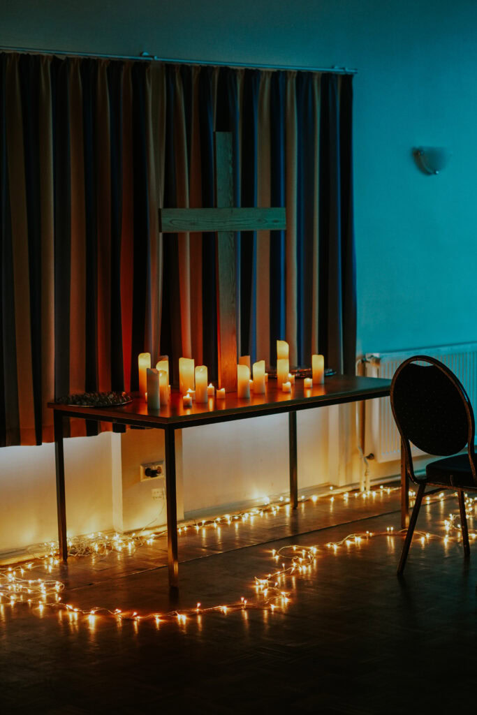 Kerzen auf einem Tisch vor einem Kreuz, mit Lichterkette am Boden.
