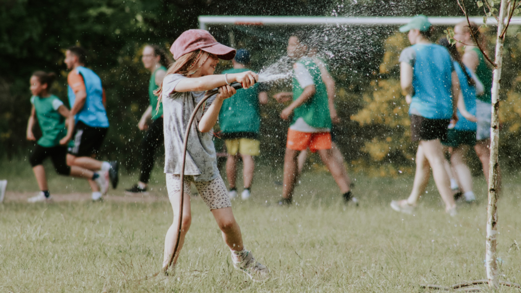 Kind spielt mit Gartenschlauch, Wasser spritzt, Kinder im Hintergrund laufen.