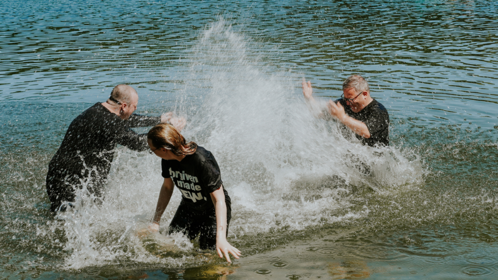 Drei Menschen spielen im Wasser, einer fällt ins Wasser, Wasser spritzt.