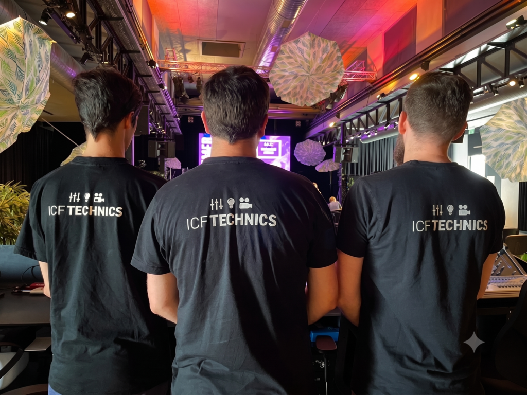 Drei Personen mit schwarzen T-Shirts mit 'ICF TECHNICS' auf dem Rücken, Blick auf die Bühne.