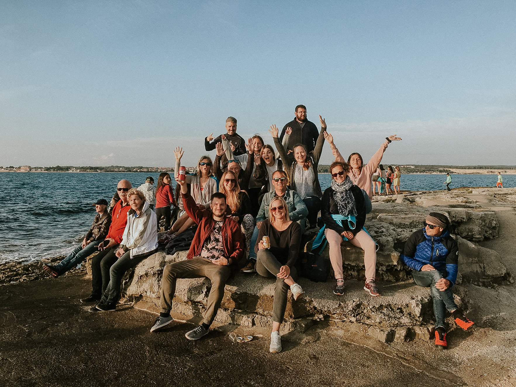 Mehrere Menschen sitzen und stehen auf Felsen am Wasser, einige winken.