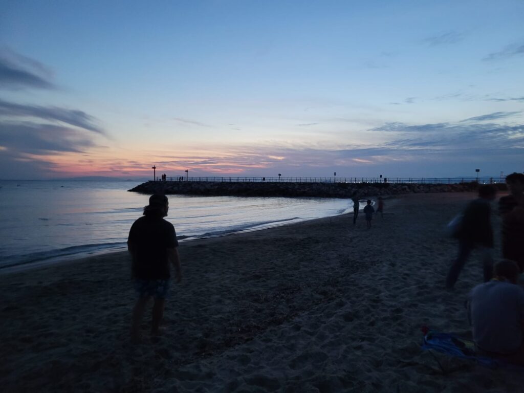 Menschen am strand bei Sonnenuntergang, Blick auf den Hafen mit Leuchttürmen.