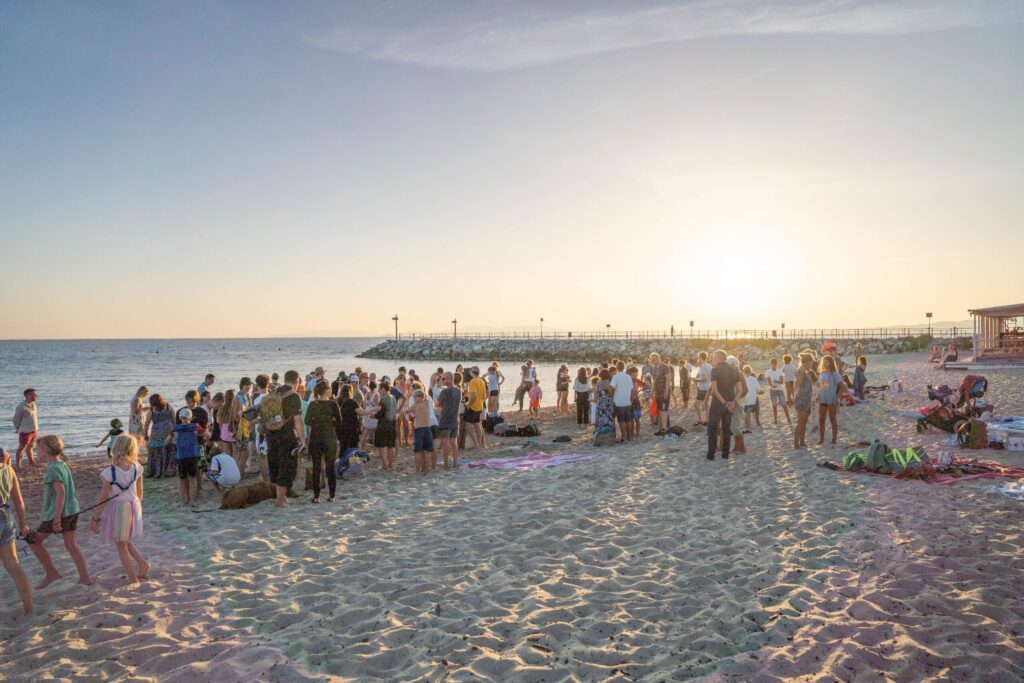 Menschenmenge am Strand bei Sonnenuntergang, einige sitzen, andere stehen.