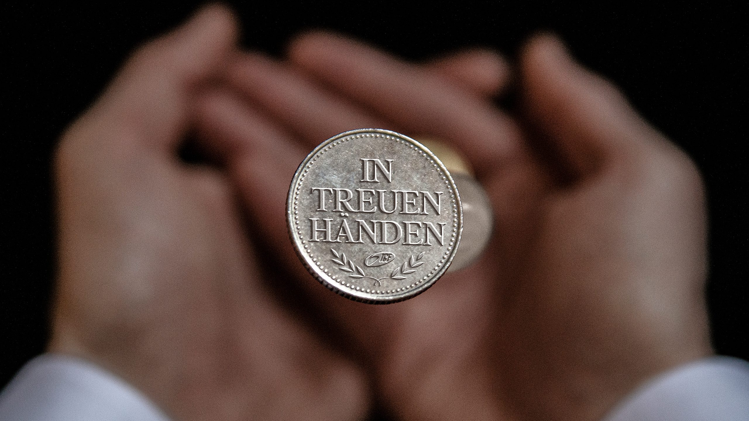Zwei Hände halten eine Münze mit der Aufschrift 'In Treuen Händen'.