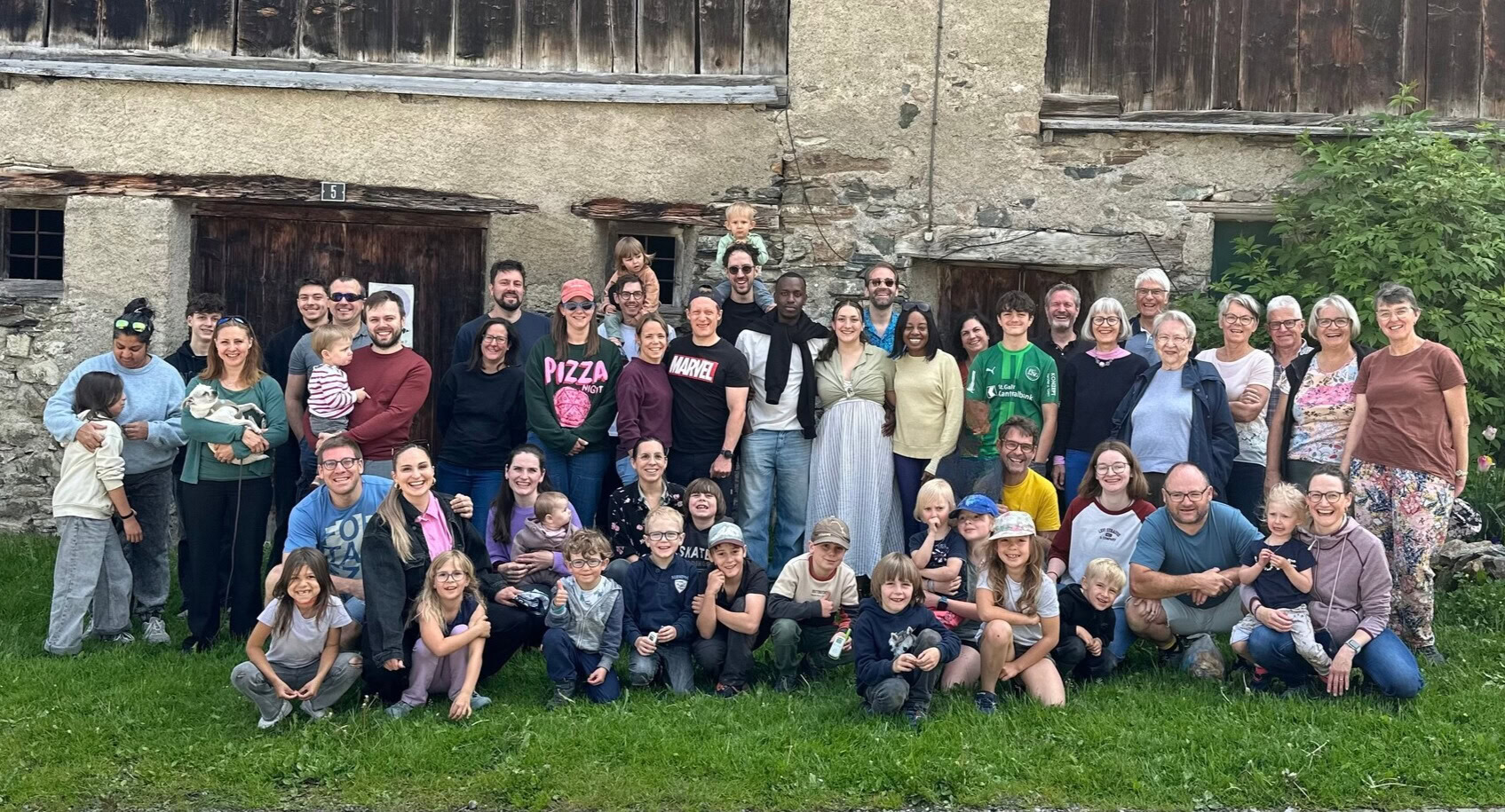 Alt Text: Fröhliche Gruppe von Menschen unterschiedlichen Alters vor einer historischen Steinmauer und Holztüren, bei einer Gemeinschaftsaktivität von ICF Zürich Oberland.