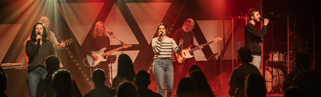 Fünf Musiker auf einer Bühne, singen und spielen Gitarre, Keyboard und Schlagzeug.