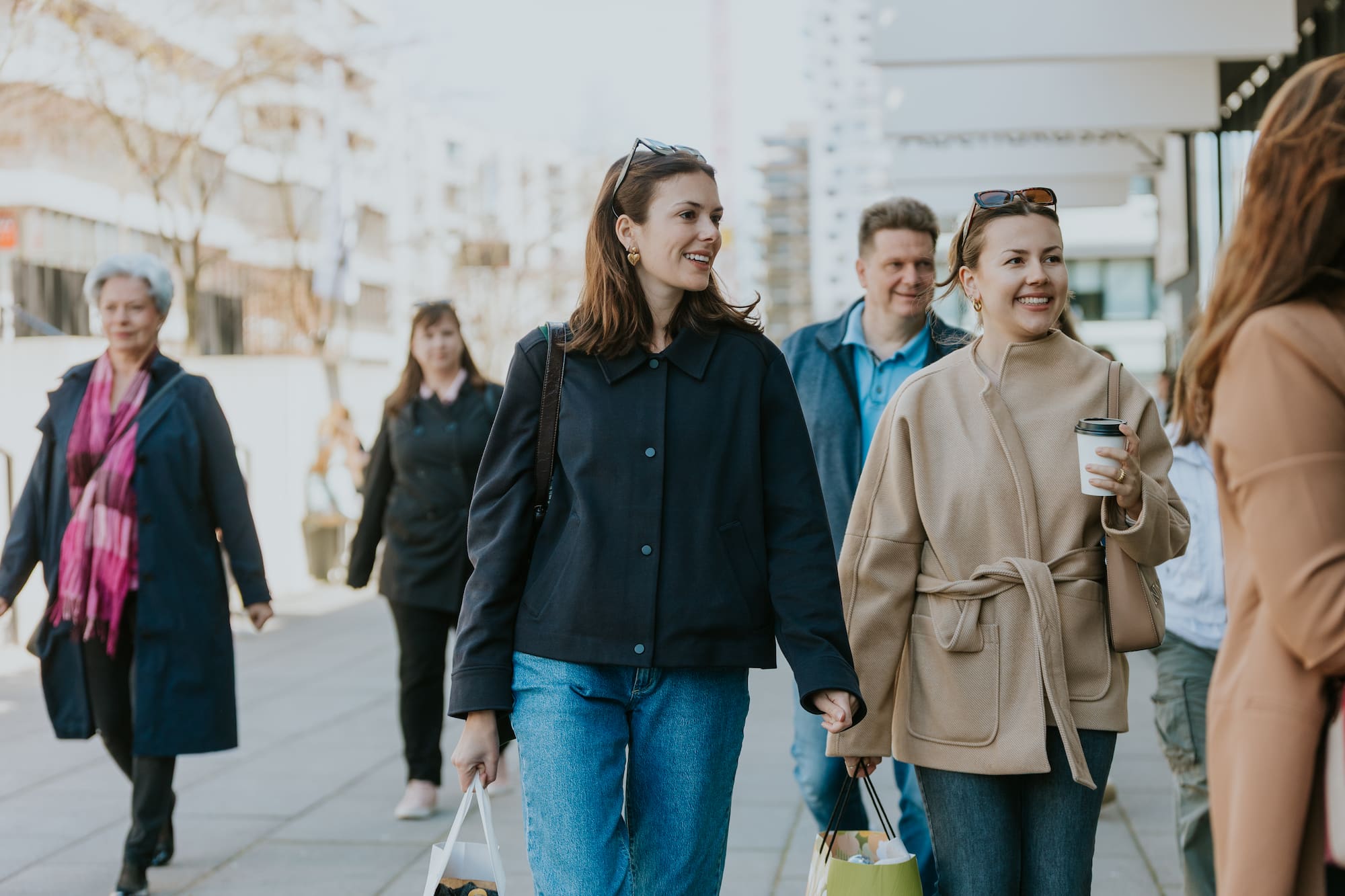 Menschen gehen auf der Straße, zwei Frauen im Vordergrund, eine hält eine Tasse, die andere trägt Ei.