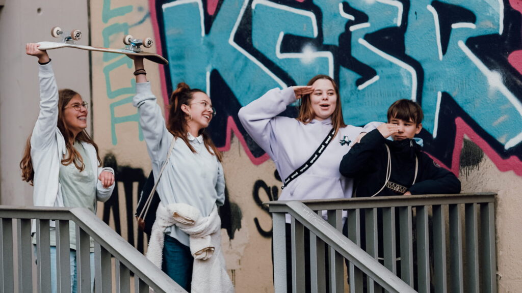 Four teenagers are standing on a staircase in front of a graffiti wall; one is holding a skateboard.