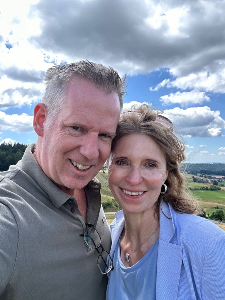 Ein Mann und eine Frau stehen im Freien vor einer Landschaft mit Wolken am Himmel.