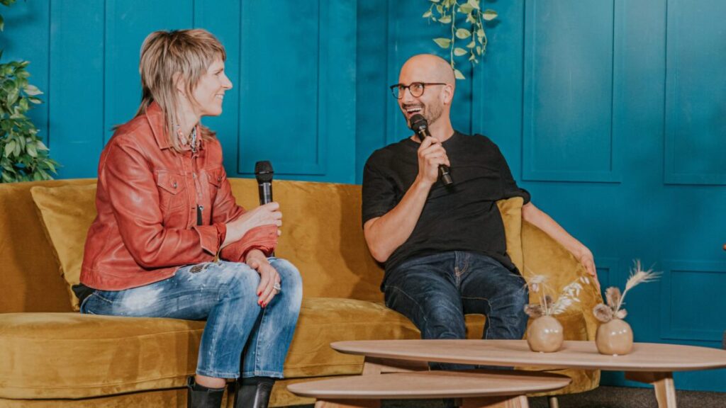 Zwei Menschen sitzen auf einer Couch und sprechen mit Mikrofonen.