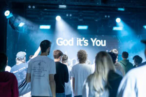 Menschen vor einer Bühne mit einem Bildschirm, der 'God it's You' zeigt.