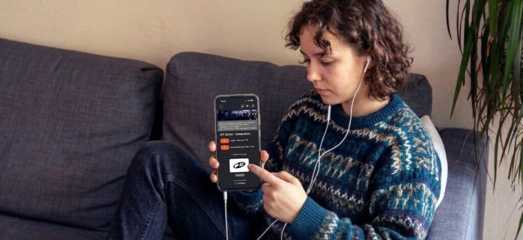 Junge Frau sitzt auf einem Sofa, hält ein Smartphone in der Hand, trägt Kopfhörer.